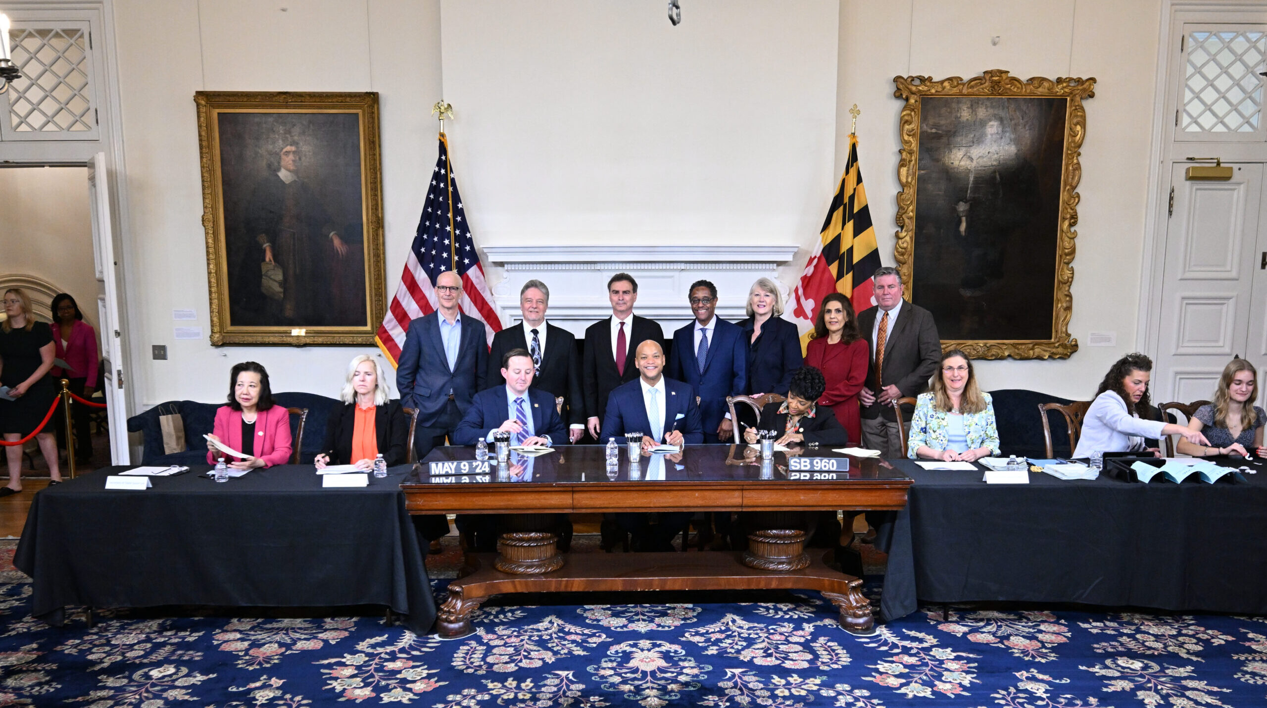 A group of officials and representatives are seated and standing around a large wooden table in a formal room. The backdrop features two large portraits and the Maryland state flag. The individuals are engaged in a meeting, with some taking notes and others looking towards the camera
