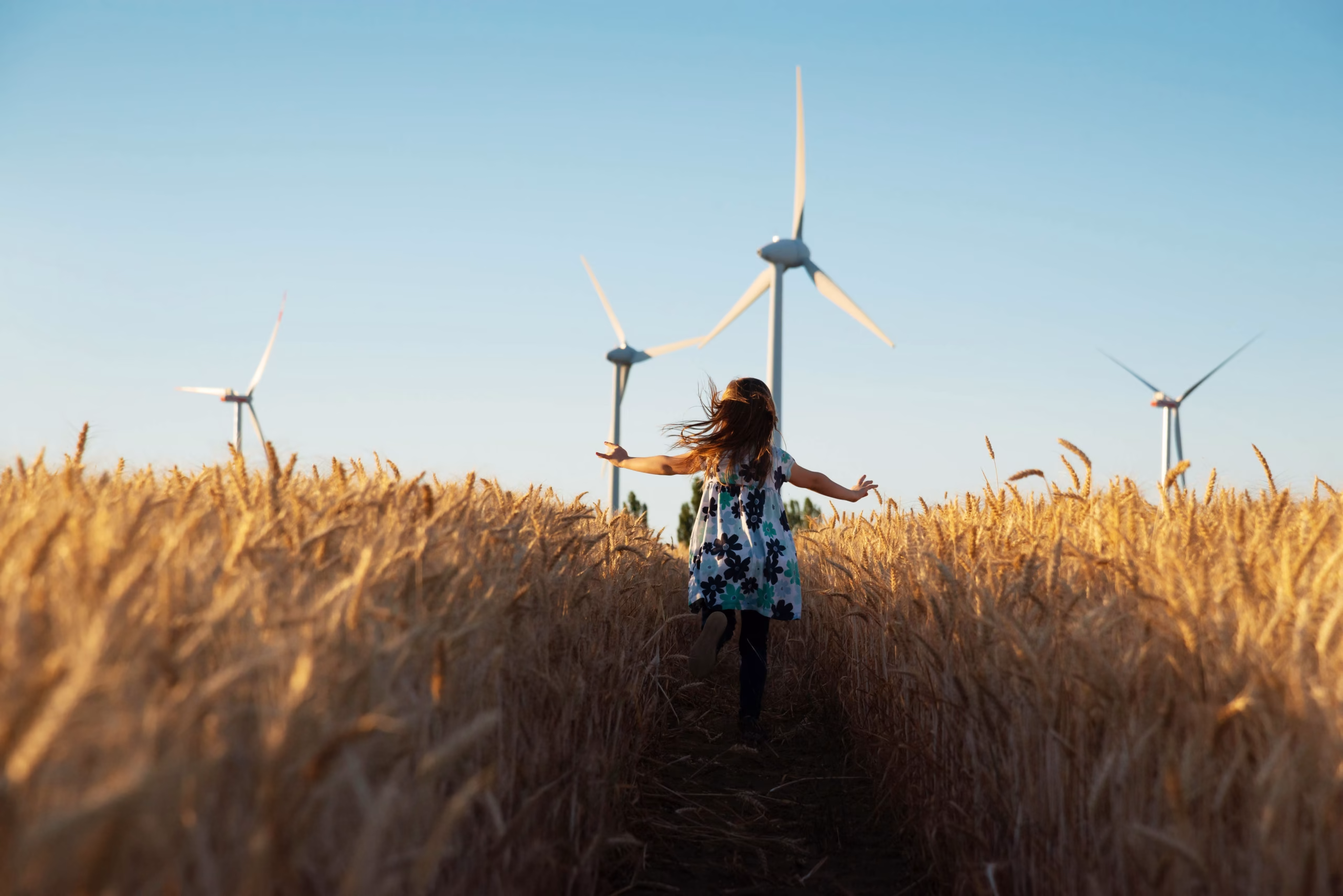 A child wearing a floral dress runs through a golden wheat field, with wind turbines visible in the background against a clear blue sky