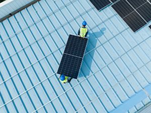 Two workers in blue helmets and yellow vests are carrying a solar panel across a blue metal roof. Several solar panels are already installed on the roof