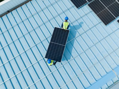 Two workers in blue helmets and yellow vests are carrying a solar panel across a blue metal roof. Several solar panels are already installed on the roof