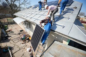 A group of workers installs solar panels on a sloped metal roof. One worker is climbing a ladder while another is securing a solar panel. Tools and equipment are scattered on the ground nearby, and the setting appears to be a residential area with trees in the background