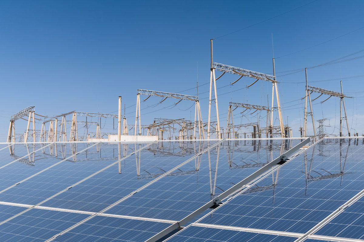A row of solar panels reflects the blue sky, with electrical transmission towers and power lines visible in the background. The scene illustrates solar panels adjacent to a power grid.