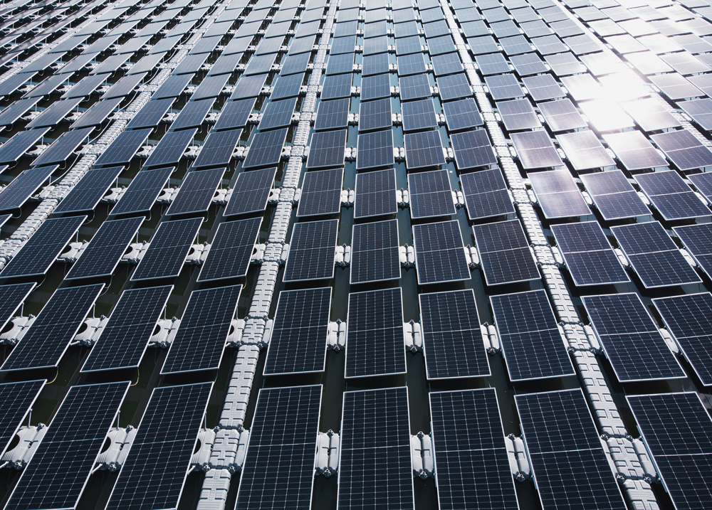 A large array of solar panels arranged in a grid pattern, reflecting sunlight. The panels cover a wide area, showcasing their uniform design and alignment
