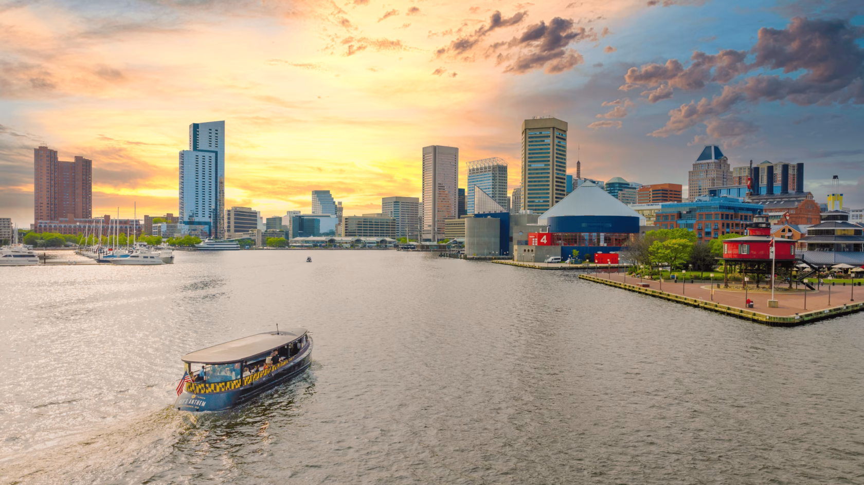 A boat navigates the water in the foreground, with a city skyline featuring tall buildings and a colorful waterfront in the background. The sky is filled with clouds and a warm sunset glow