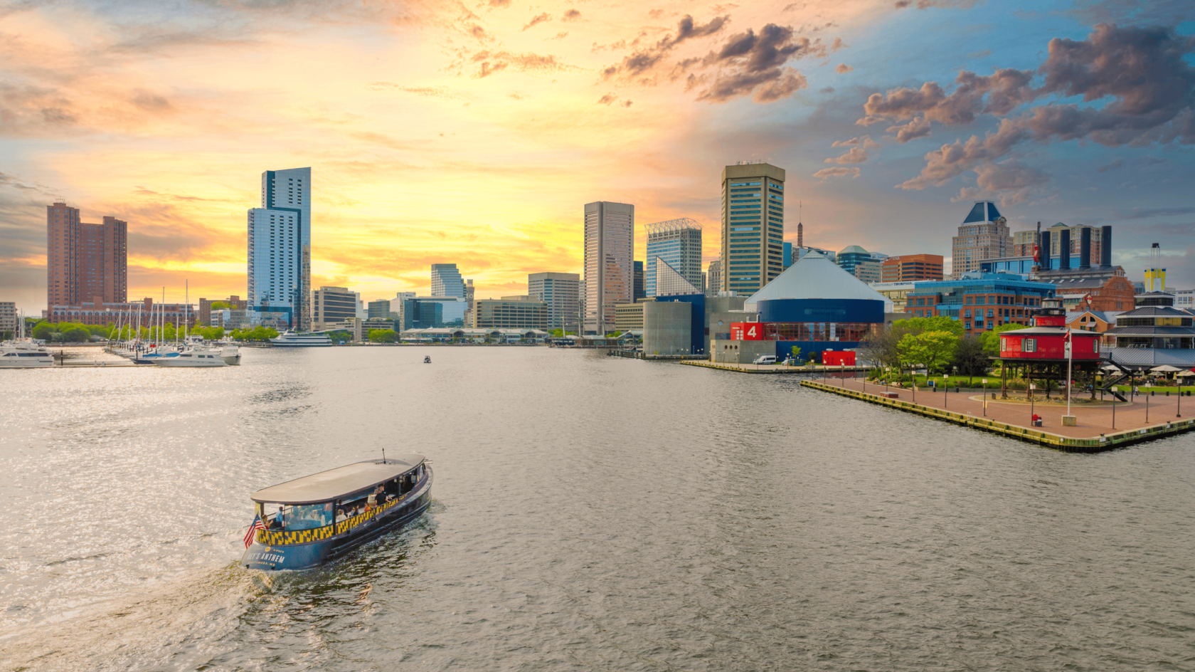A boat navigates the water in the foreground, with a city skyline featuring tall buildings and a colorful waterfront in the background. The sky is filled with clouds and a warm sunset glow