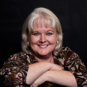 A woman with shoulder-length blonde hair smiles while resting her arms on a surface. She is wearing a patterned top and has earrings. The background is dark