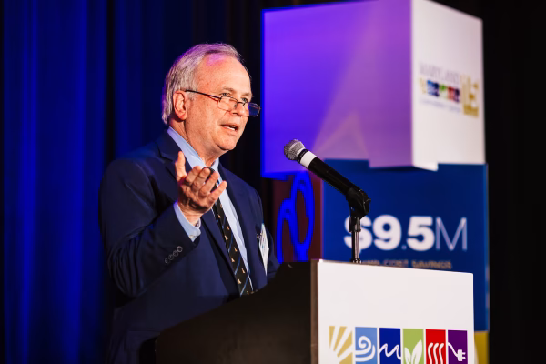A man in a blue suit speaks at a podium with a microphone, gesturing with his right hand. Behind him, colorful banners display various logos and the amount "$9.5M." The setting appears to be a formal event or conference