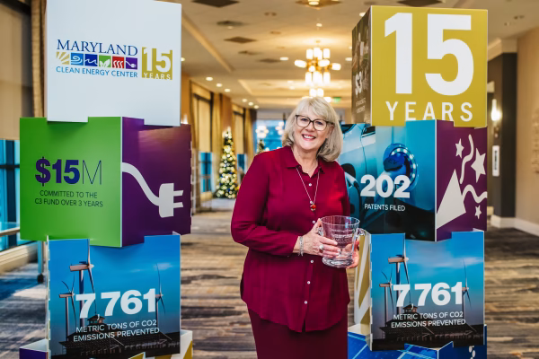 A woman with blonde hair and glasses stands holding a clear award in front of two large display boards. The boards highlight the Maryland Clean Energy Center's 15-year anniversary.