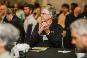 A man with glasses and gray hair sits at a table, clapping his hands. He is wearing a black LG-branded shirt and appears to be engaged in a conference or event, surrounded by other attendees in a blurred background
