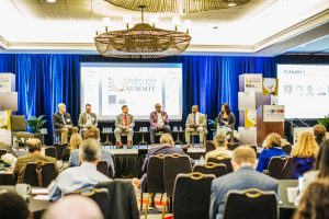 A panel discussion taking place at the Maryland Summit, featuring six speakers seated on a stage with a blue backdrop. The audience is visible in the foreground, facing the panel. The setting includes a large chandelier and various event branding elements