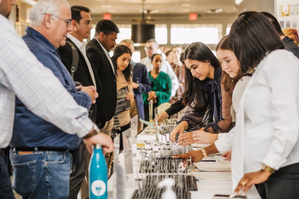 A diverse group of people is gathered around a table displaying various products. Some individuals are examining items, while others are engaged in conversation. The setting appears to be a networking or promotional event, with a bright and open atmosphere