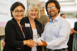 A woman in a black blazer shakes hands with another woman in a gray blazer, while a man in a light blue shirt holds an award or plaque. All three are smiling and appear to be at a formal event