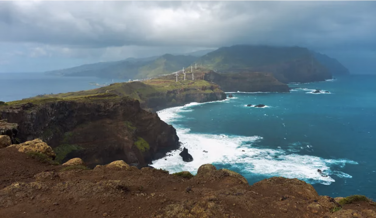 A coastal landscape featuring rugged cliffs and crashing waves, with a mountainous backdrop. Wind turbines are visible on the hills in the distance, under a cloudy sky