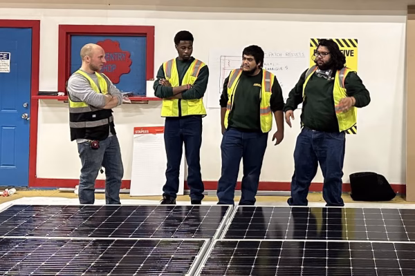 A group of four individuals stands near a large array of solar panels indoors. They are wearing safety vests and engaged in discussion. Two individuals are facing each other, while the other two are observing. The background features a blue door and various posters on the walls