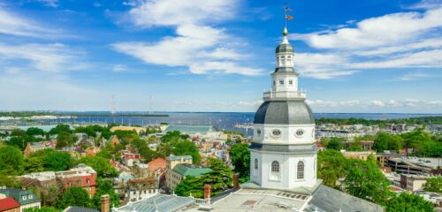 A view of a city with a prominent dome structure. The dome is part of a historic building, likely a government or civic structure. In the background, there is a body of water with boats and a clear blue sky.