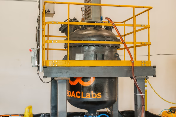A large industrial mixing tank with a yellow safety railing, located in a facility. The tank is equipped with a blue motor on top and has pipes connected to it. A blue valve is visible at the bottom, and the area is surrounded by a black containment barrier