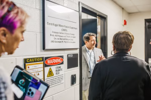 A group of people stands in a hallway outside a door labeled "Centrifugal Mirror Fusion Experiment (CMFX)" with a sign indicating the professor's name and affiliation.