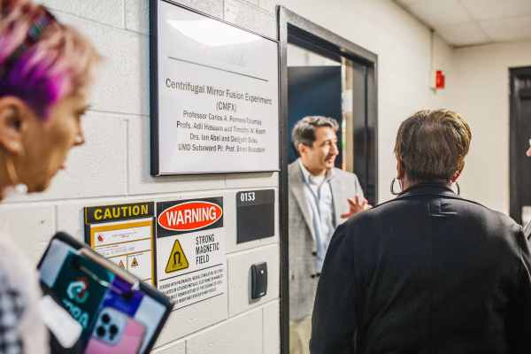 A group of people stands in a hallway outside a door labeled "Centrifugal Mirror Fusion Experiment (CMFX)" with a sign indicating the professor's name and affiliation.
