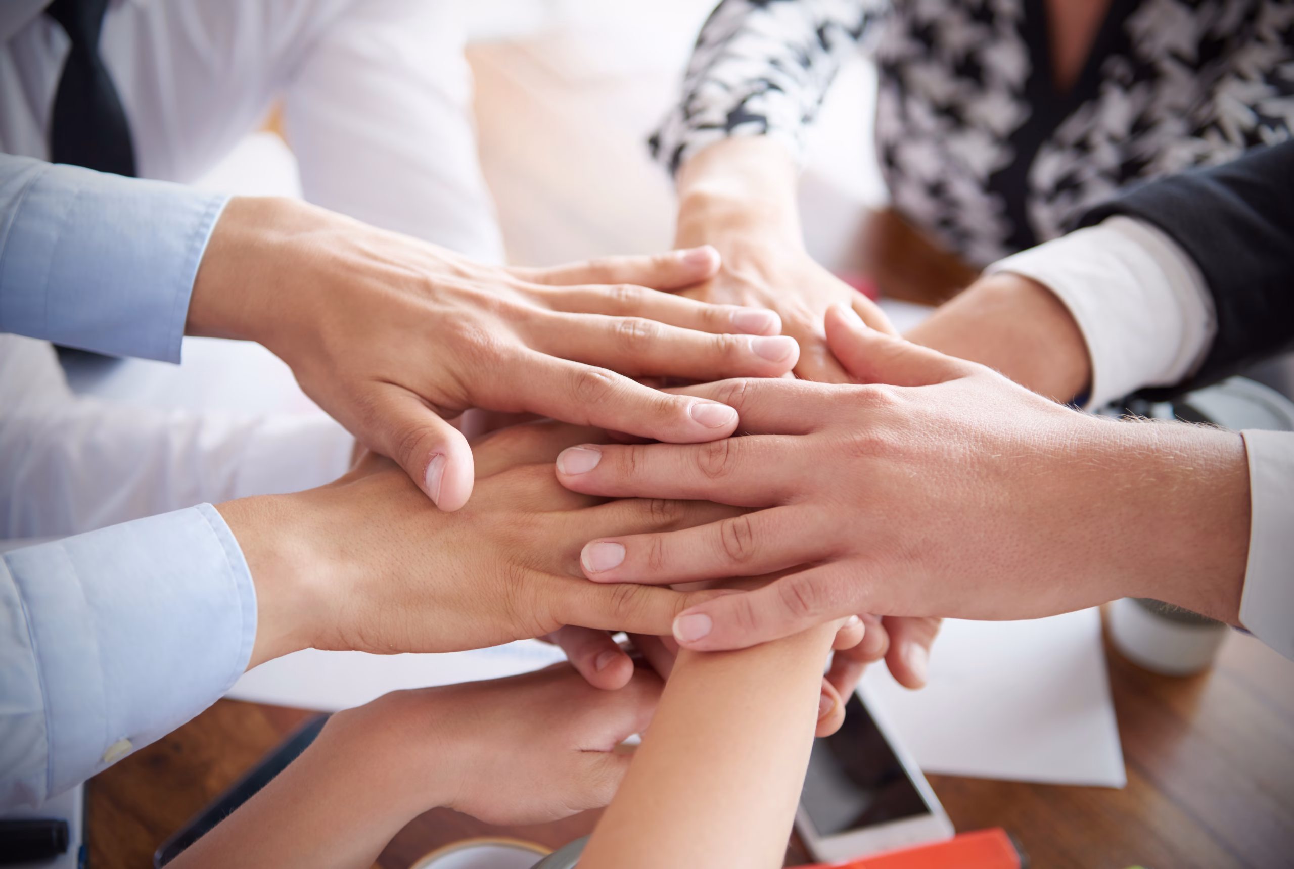 A group of hands from multiple people stacked together on a wooden table, symbolizing teamwork and collaboration. The hands vary in skin tone and are positioned in a circular arrangement