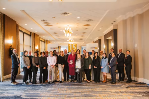 A group of approximately 20 people stands together in a well-lit hotel conference room, smiling and posing for a photo. They are dressed in business casual attire and are arranged in two rows. A large banner in the background displays the number "15