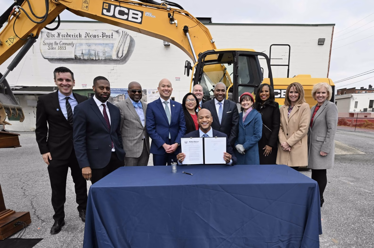 A group of officials and community members stands behind a table with a document, celebrating a groundbreaking event. In the background, a large yellow excavator is visible. The individuals are dressed in formal attire, and some are smiling at the camera