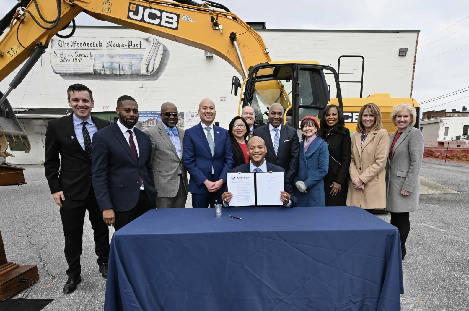 A group of officials and community members stands behind a table with a document, celebrating a groundbreaking event. In the background, a large yellow excavator is visible. The individuals are dressed in formal attire, and some are smiling at the camera
