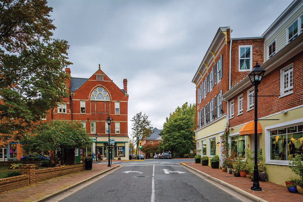 A view of a tree-lined street featuring brick buildings on either side. On the left, a historic building with a pointed roof and decorative windows. On the right, a modern building with large windows and an orange awning. The street is empty, and the sky is overcast