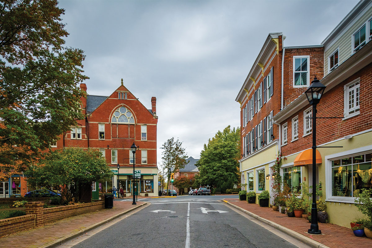 A view of a tree-lined street featuring brick buildings on either side. On the left, a historic building with a pointed roof and decorative windows. On the right, a modern building with large windows and an orange awning. The street is empty, and the sky is overcast