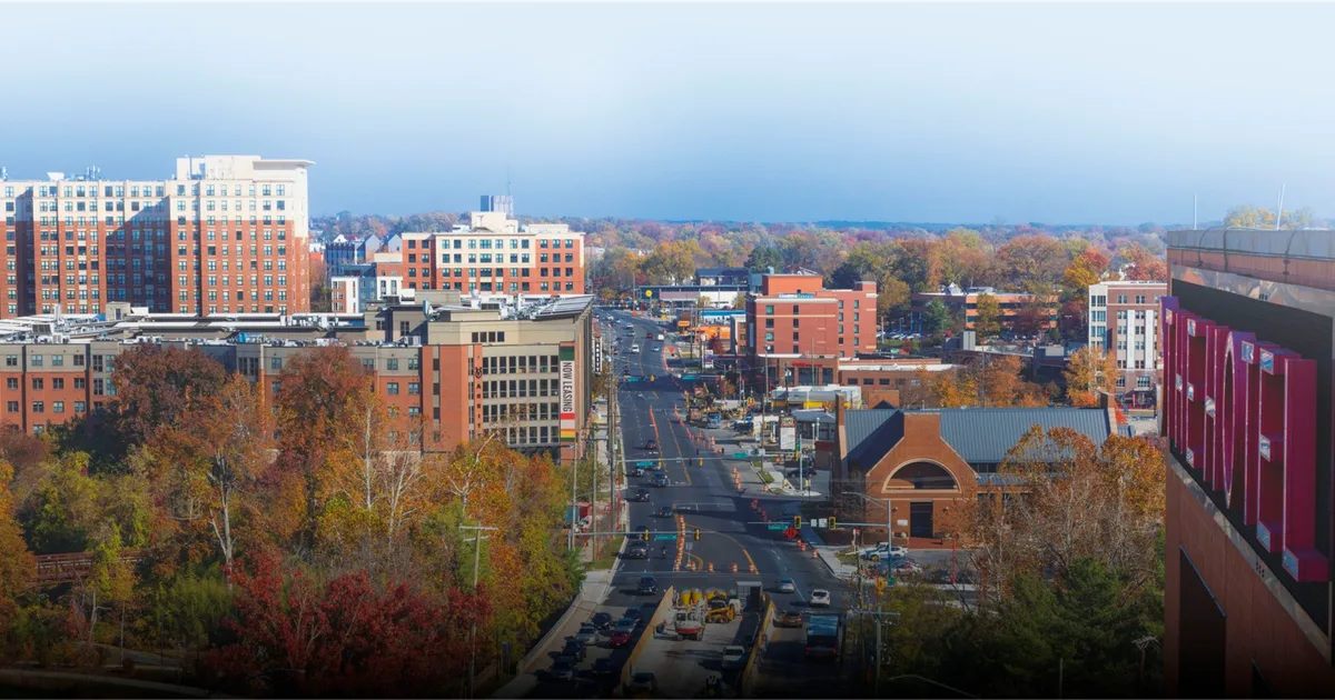 A view of a cityscape featuring a mix of modern and older buildings, with a tree-lined street running through the center. The scene shows autumn foliage in various colors, indicating the season. In the background, several high-rise buildings and a clear sky are visible
