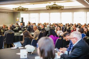 A large audience is seated in a conference room, attentively listening to a speaker at the front. The attendees are a diverse group, with some taking notes or using devices. Tables are set with notebooks and small floral arrangements. Large windows provide natural light to the space