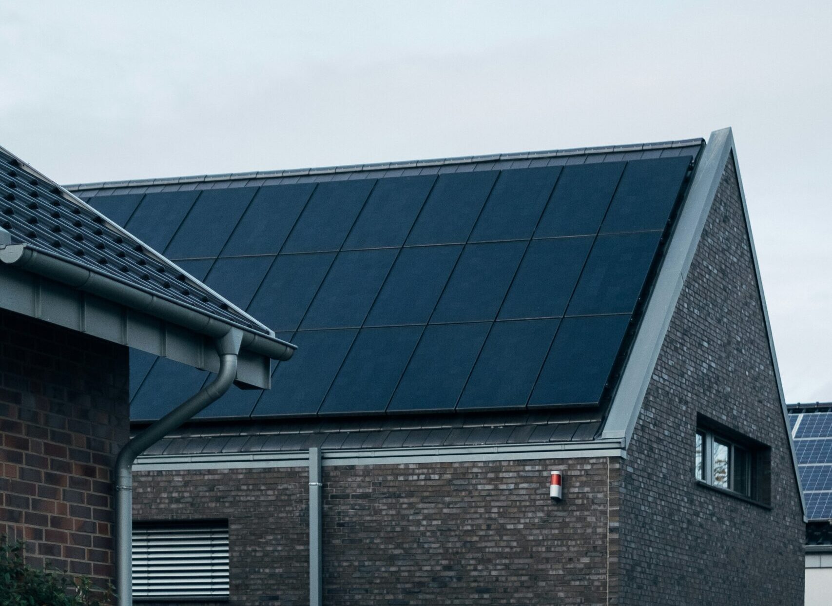 A sloped roof of a modern brick house features dark solar panels. Nearby, another building with a gray roof is visible. The sky is overcast