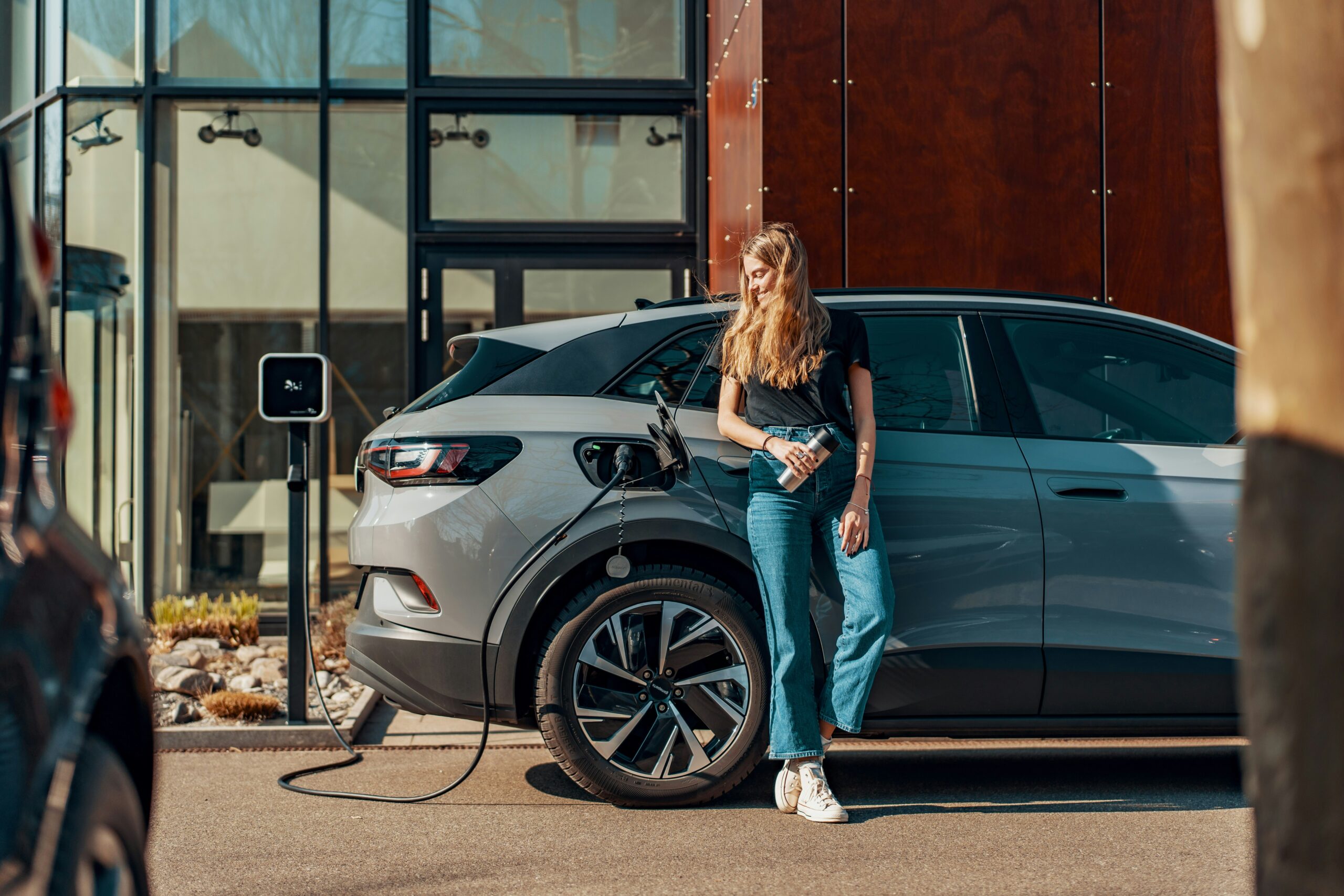 A woman with long hair stands next to a gray electric vehicle that is plugged into a charging station. She is wearing a black t-shirt and blue jeans, holding a drink in one hand. The background features a modern building with large windows