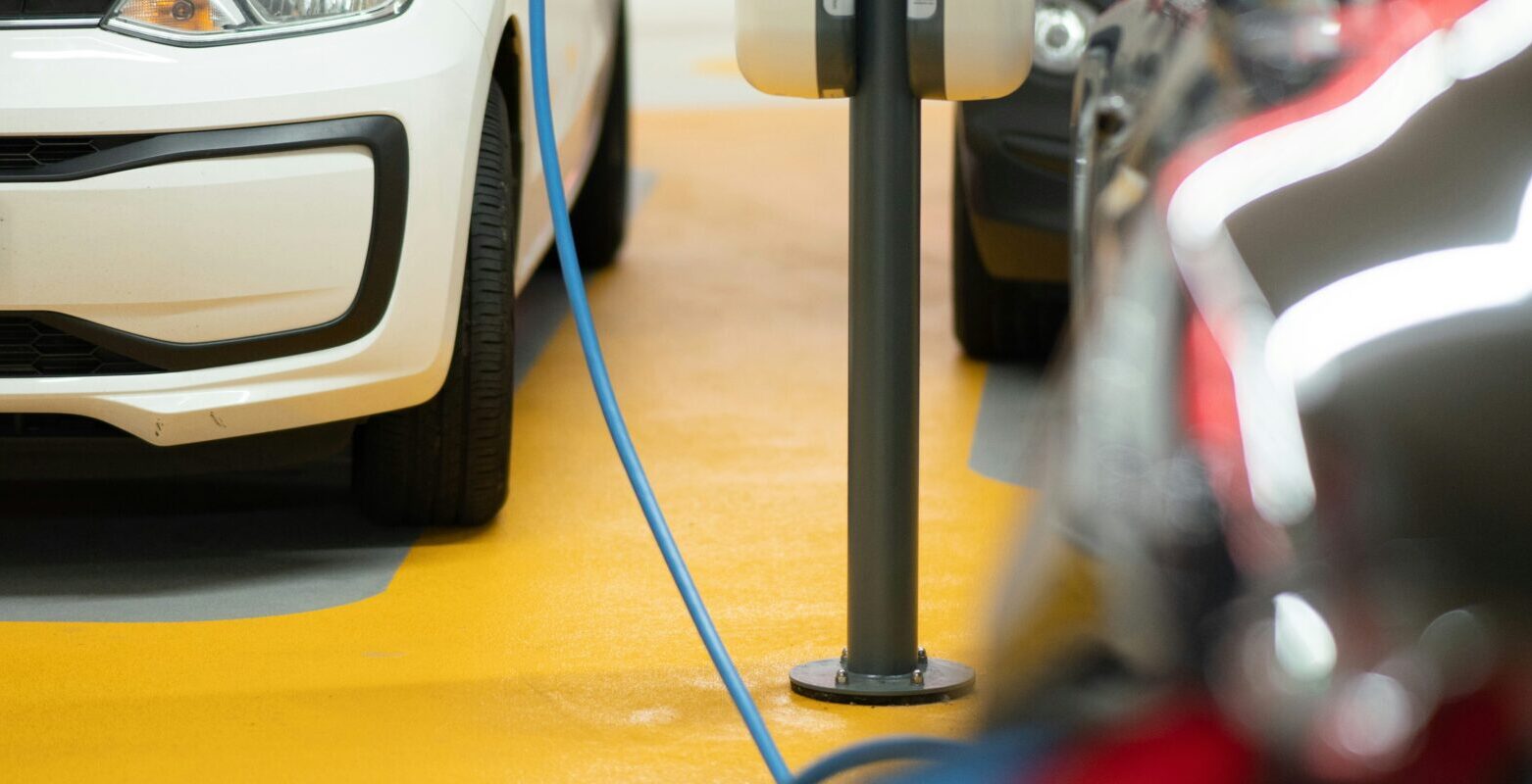 A white electric car is plugged into a charging station in a parking garage. The charging station is mounted on a pole, with blue cables connecting it to the vehicle. Other cars are parked nearby in the background