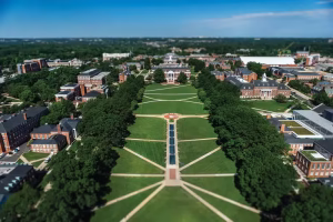 Aerial view of a university campus featuring a large, open green space with symmetrical pathways and trees. Surrounding buildings include academic structures and residential halls, with a clear blue sky in the background