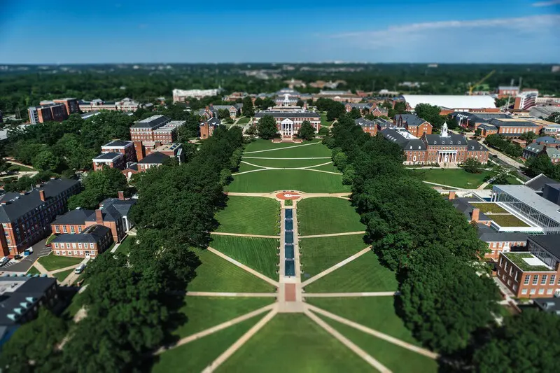 Aerial view of a university campus featuring a large, open green space with symmetrical pathways and trees. Surrounding buildings include academic structures and residential halls, with a clear blue sky in the background