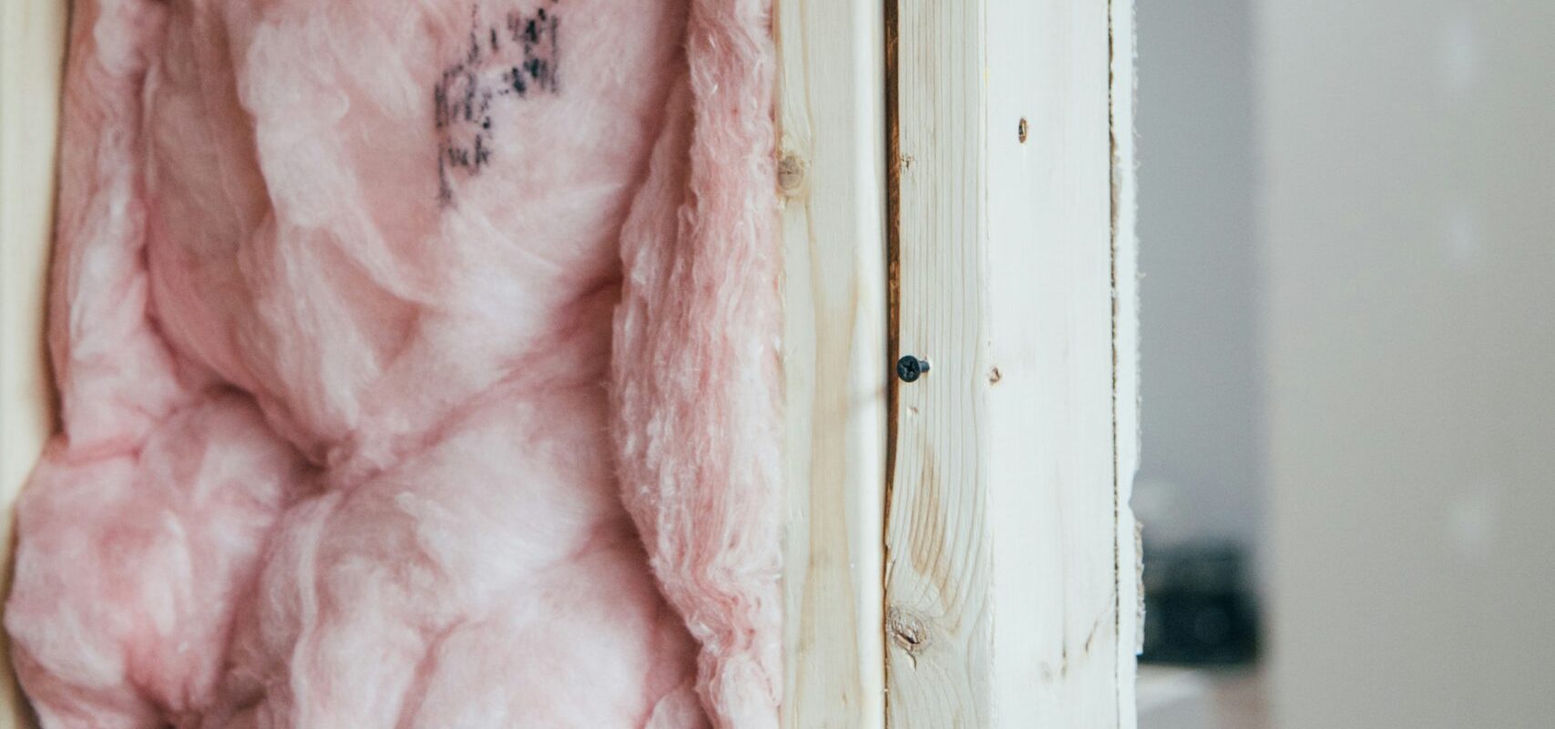 Insulation material made of pink fiberglass is installed between wooden framing in a wall. The surrounding area shows unfinished drywall and construction debris on the floor