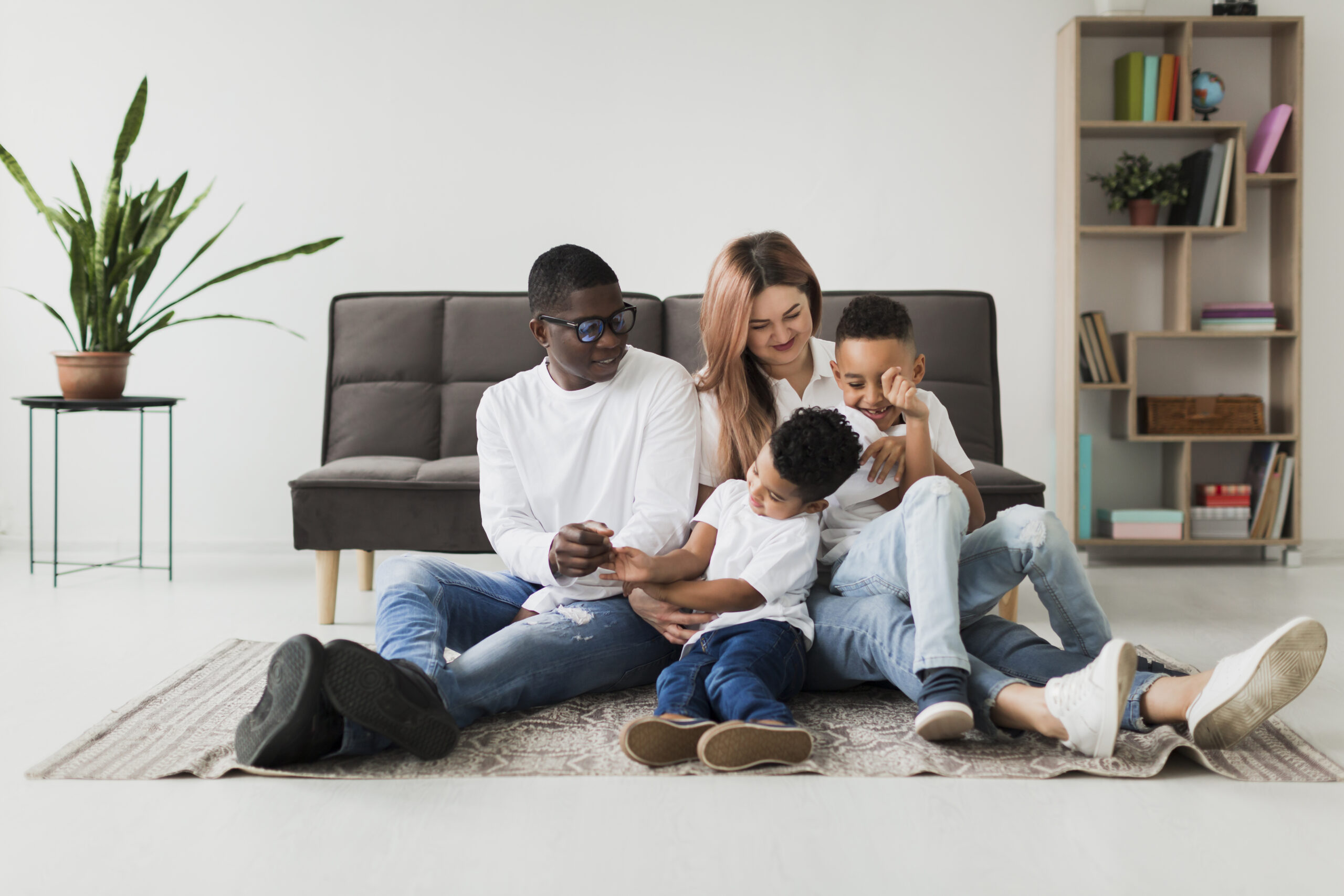 A family of four sits together on a rug in a bright room. Two adults, a man wearing sunglasses and a woman with long hair, are interacting with two young boys. The boys are playfully engaged with each other, while the adults smile at them. A potted plant and a bookshelf are visible in the background