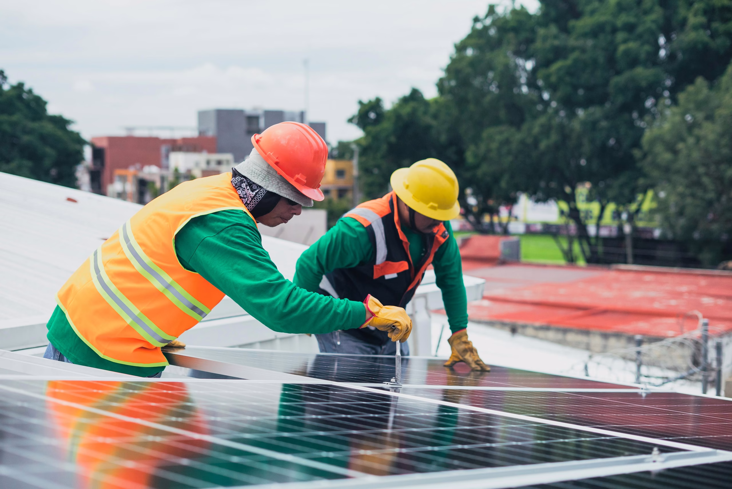 Two construction workers are installing solar panels on a rooftop. One worker, wearing a yellow hard hat and orange safety vest, is adjusting a panel, while the other, in a gray hard hat and green shirt, is securing another panel. The background shows a cityscape with buildings and trees