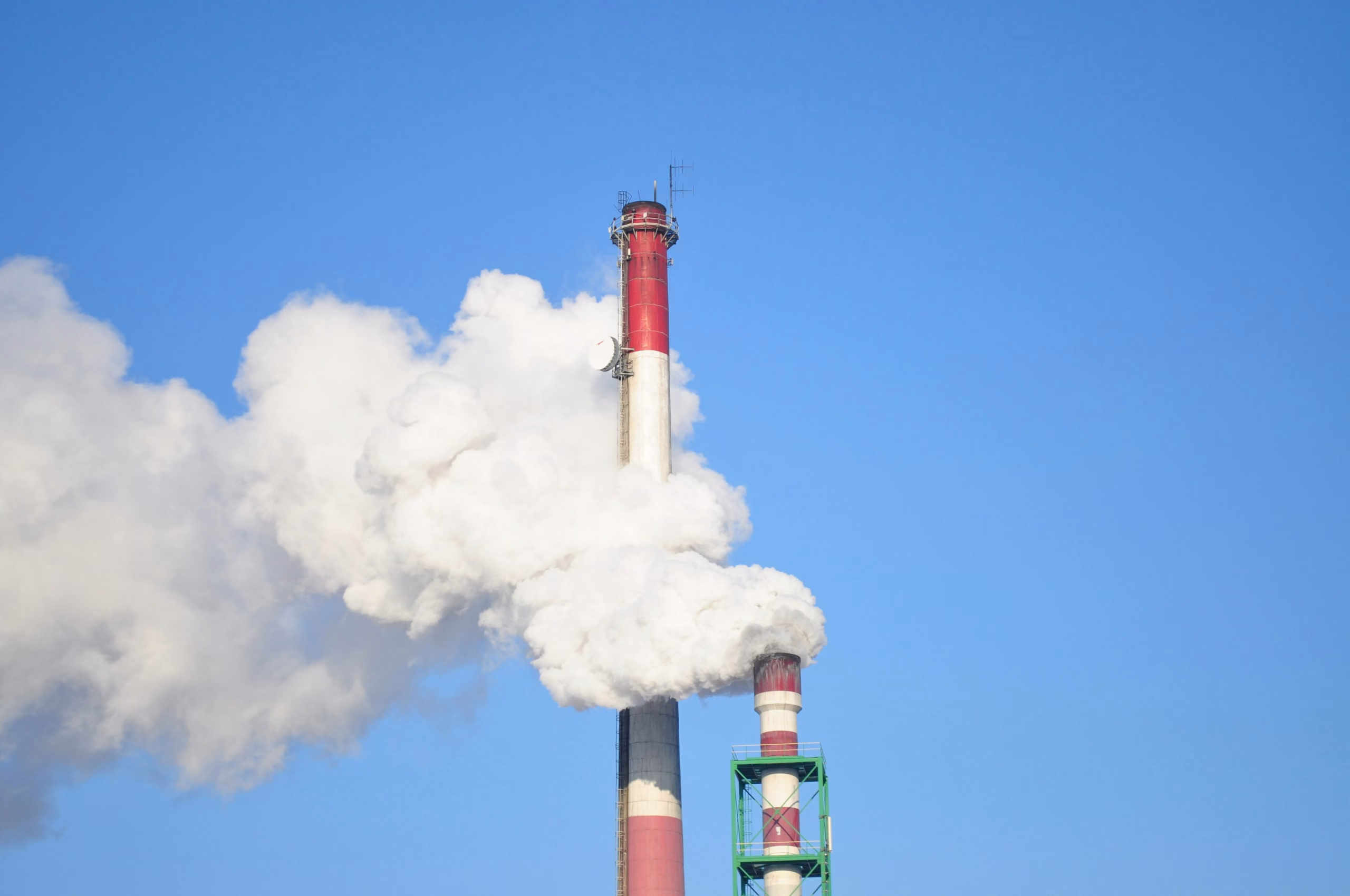 A tall industrial smokestack emits thick white smoke against a clear blue sky. The smokestack is painted with red and white stripes and is part of a larger facility