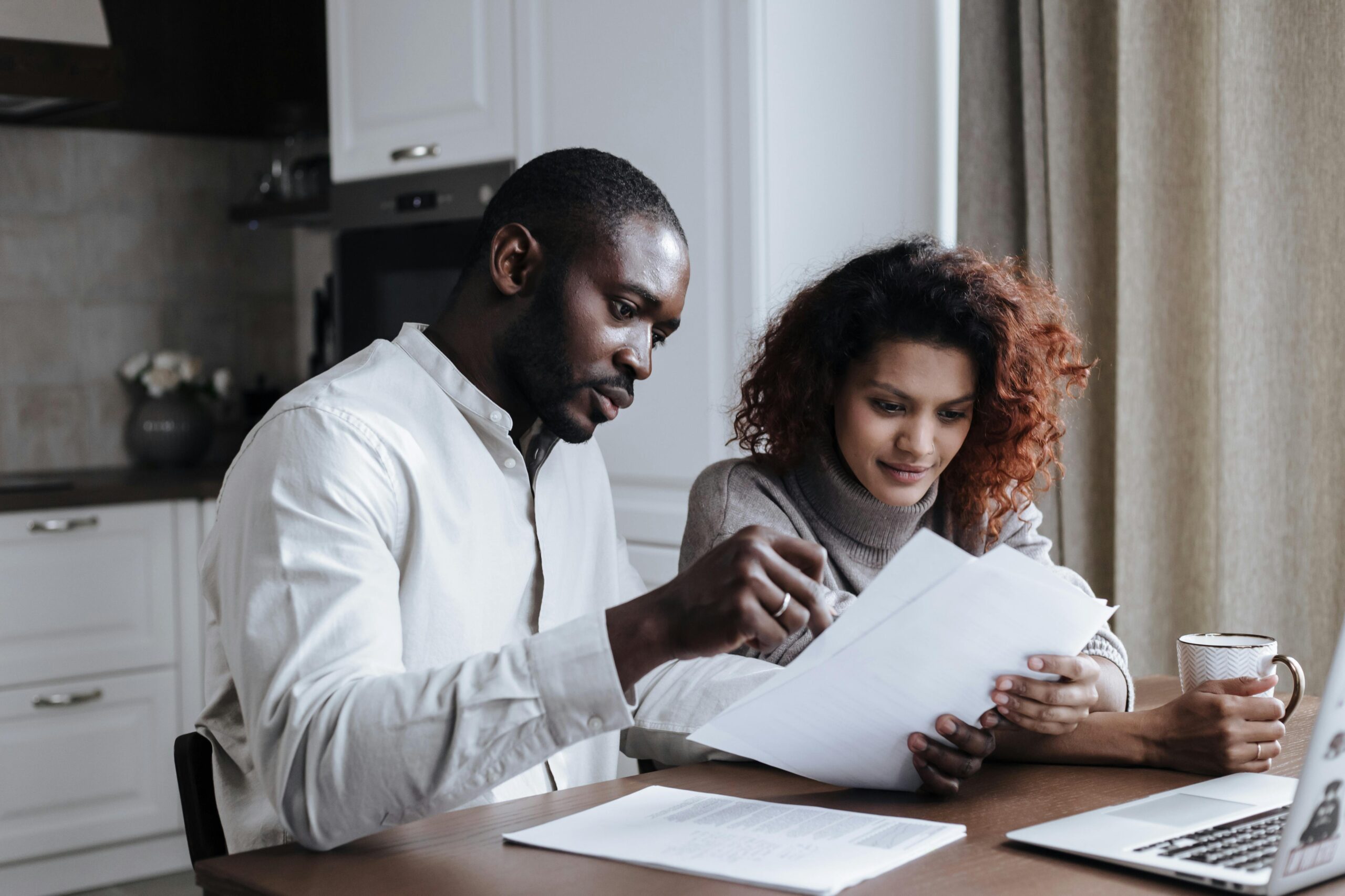 A man and a woman sit at a table, reviewing documents together. The man, wearing a light-colored shirt, points at the papers, while the woman, in a sweater, holds a cup and looks at the documents. A laptop is open on the table