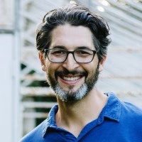 A man with dark, wavy hair and a beard smiles while wearing glasses and a blue shirt. He is outdoors, with a blurred background of an industrial park.