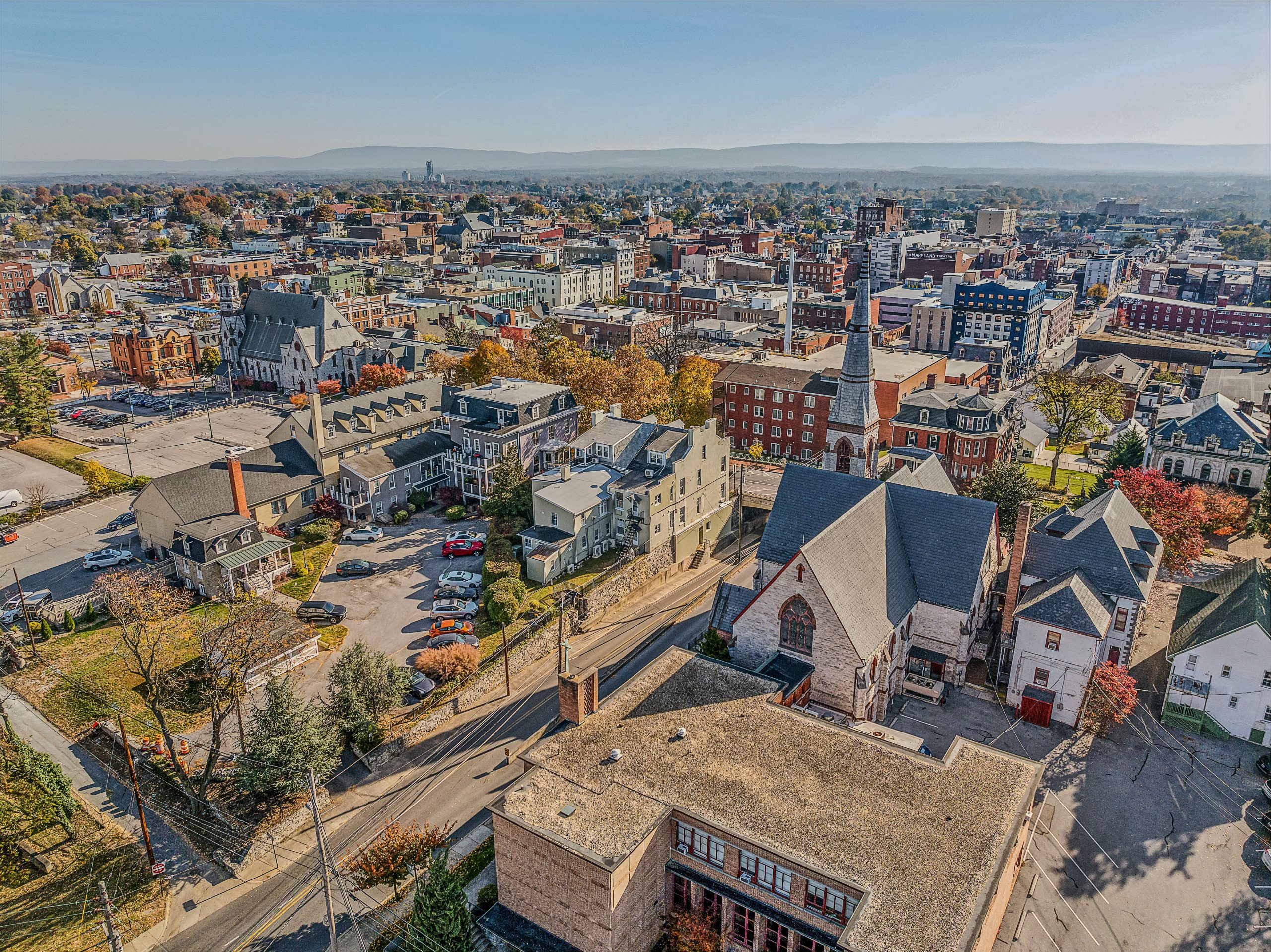 Aerial view of a cityscape featuring a mix of residential and commercial buildings, with several churches visible. The scene includes tree-lined streets and a backdrop of distant mountains under a clear blue sky