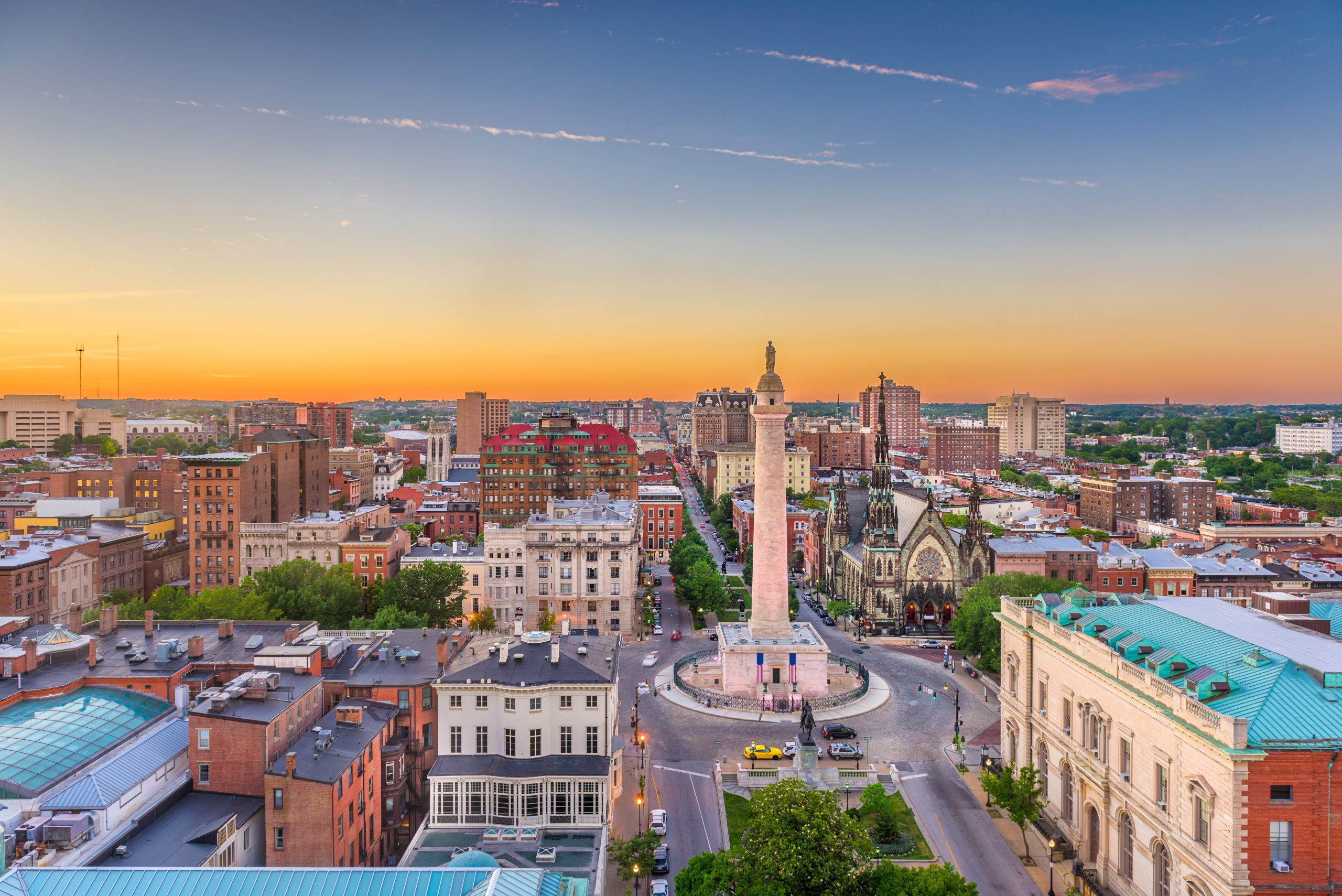 A city skyline at sunset featuring a prominent monument in a circular plaza. Surrounding buildings include a mix of historic and modern architecture, with trees and streets visible. The sky displays warm hues of orange and pink as the sun sets