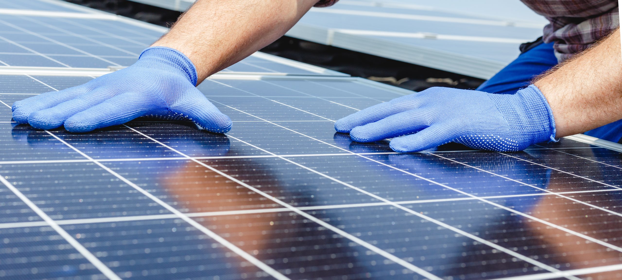A person wearing blue gloves is installing or adjusting a solar panel, with their hands placed on the surface of the panel. The background shows additional solar panels