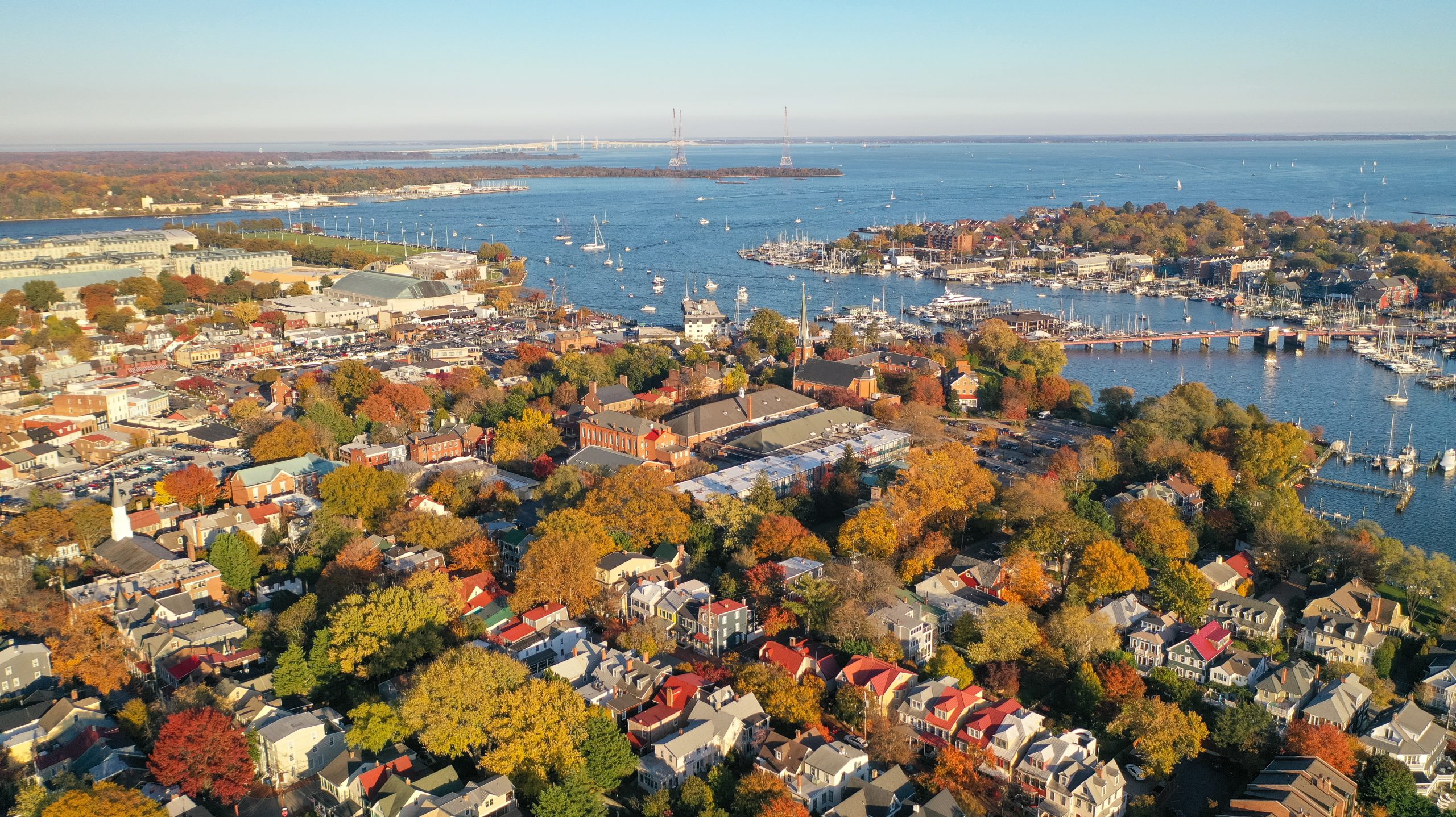Aerial view of a coastal town with a mix of residential buildings and marinas. The landscape features autumn foliage in vibrant colors, with boats docked in the water and a bridge visible in the distance.