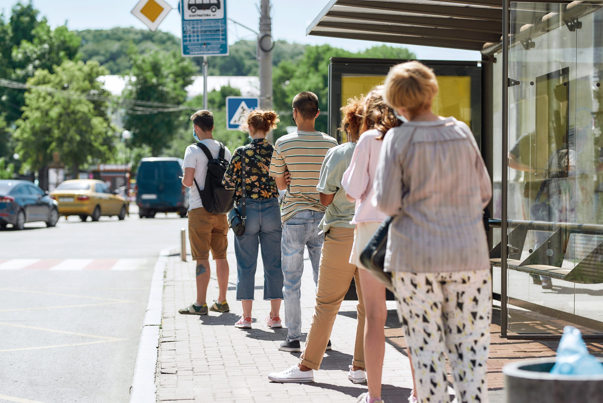 A group of seven people stands in line at a bus stop on a sunny day. They are dressed in casual summer clothing, with some wearing shorts and others in light shirts. The background features trees and vehicles, indicating a city setting