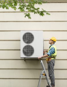 A technician in a yellow hard hat and reflective vest is standing on a ladder, installing an outdoor air conditioning unit mounted on a beige wall. The technician is focused on the unit, which has two large circular vents