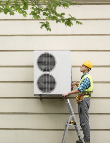 A technician in a yellow hard hat and reflective vest is standing on a ladder, installing an outdoor air conditioning unit mounted on a beige wall. The technician is focused on the unit, which has two large circular vents