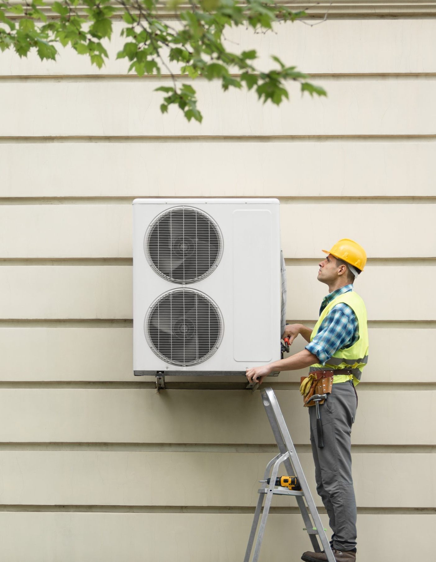 A technician in a yellow hard hat and reflective vest is standing on a ladder, installing an outdoor air conditioning unit mounted on a beige wall. The technician is focused on the unit, which has two large circular vents