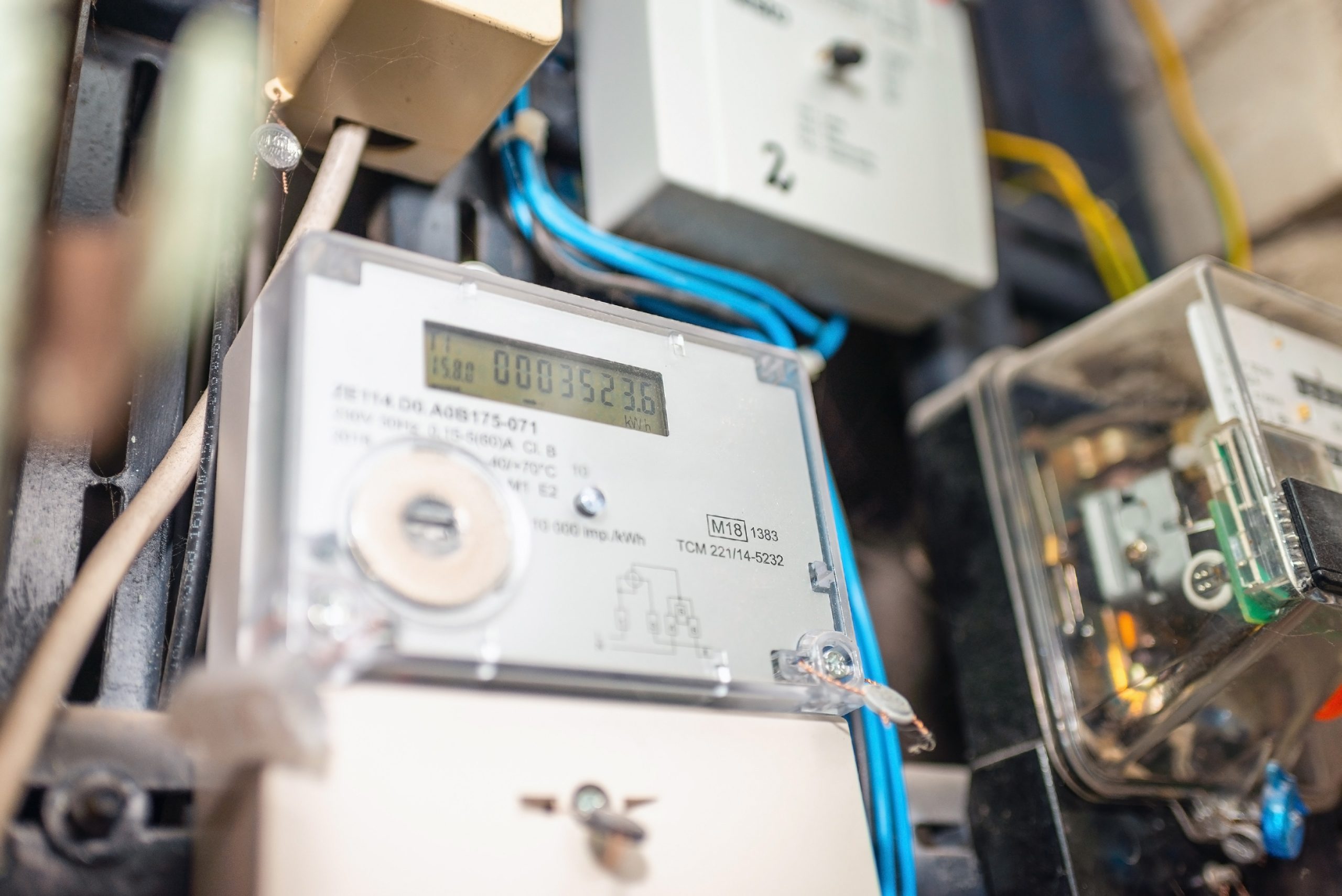 A digital electricity meter mounted on a wall, displaying a numerical reading. Wires in various colors are connected to the meter.